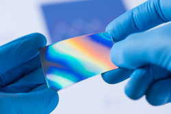 Piece of new type of material or thin coating on plastic with improved properties in laboratory in scientist hands, iridescent and pressure sensitive
